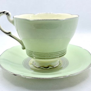 May include: A vintage teacup and saucer set with a pale green color and silver trim. The cup has a delicate, scalloped rim and the saucer has a swirling pattern.