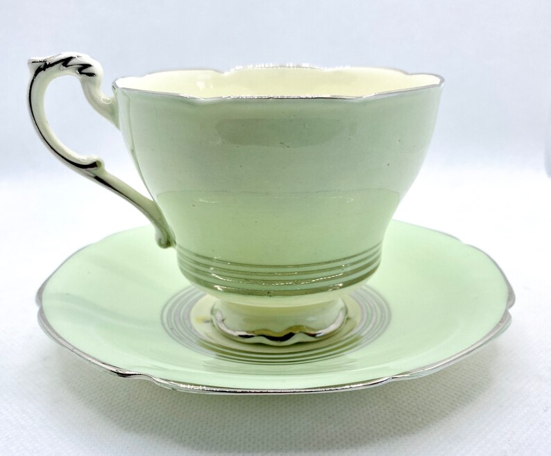 May include: A vintage teacup and saucer set with a pale green color and silver trim. The cup has a delicate, scalloped rim and the saucer has a swirling pattern.