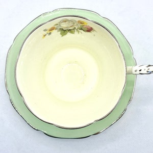 May include: A vintage teacup and saucer set with a delicate floral design. The cup and saucer are both white with a light green rim and a gold trim. The floral design features white roses and green leaves.