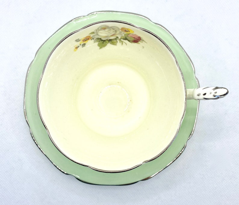 May include: A vintage teacup and saucer set with a delicate floral design. The cup and saucer are both white with a light green rim and a gold trim. The floral design features white roses and green leaves.