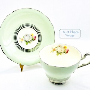 May include: A vintage teacup and saucer set with a delicate floral design. The cup and saucer are a pale green color with a gold trim. The floral design features white roses with pink and yellow accents.