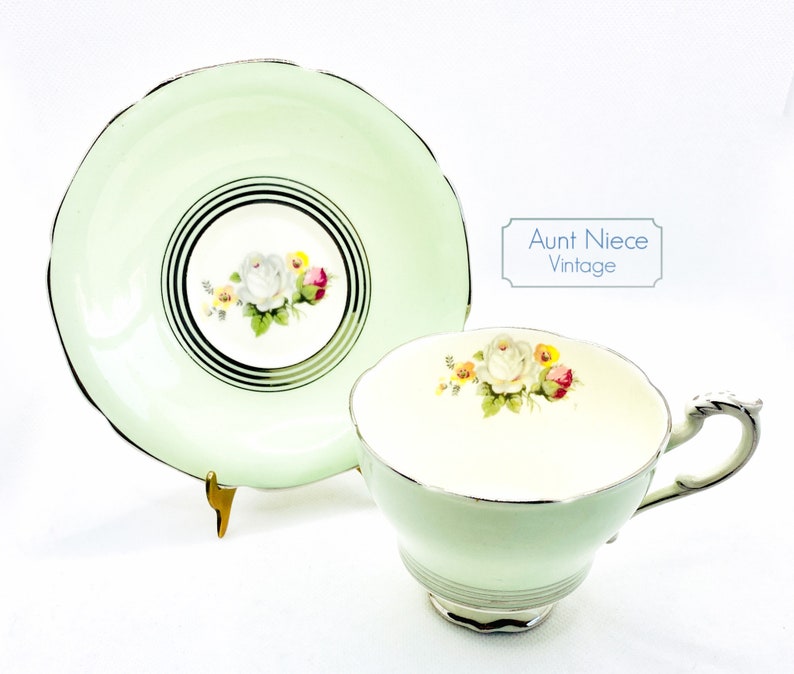 May include: A vintage teacup and saucer set with a delicate floral design. The cup and saucer are a pale green color with a gold trim. The floral design features white roses with pink and yellow accents.
