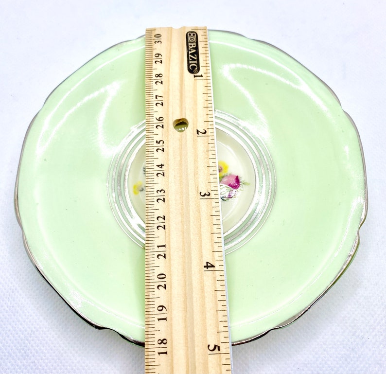 May include: A light green saucer with a silver rim and a floral design in the center. The saucer measures 5 inches in diameter.