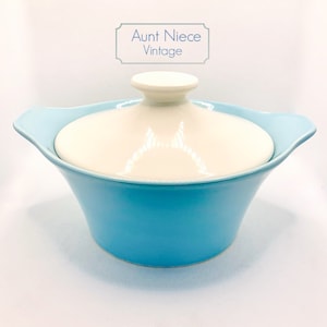 May include: A light blue ceramic casserole dish with a white lid. The dish has two handles on the sides. The lid has a knob on top. The dish is labeled "Aunt Niece Vintage".
