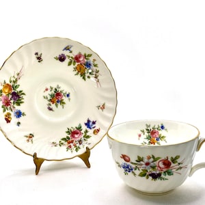 May include: A white teacup and saucer with a floral pattern. The cup and saucer have a gold trim and are decorated with a variety of colorful flowers.