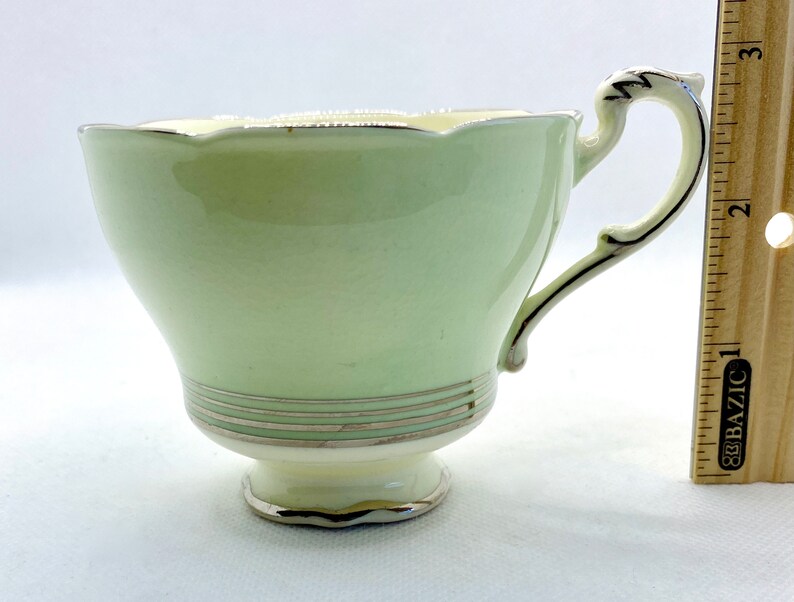 May include: A pale green teacup with a scalloped rim and a silver trim. The cup has a delicate handle and a small base.