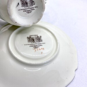 May include: A white teacup and saucer set with a gold trim. The bottom of the cup and saucer have a brown stamp that reads "Paragon by Appointment to H.M. The Queen & H.M. Queen Mary Fine Bone China England Reg'd". The saucer also has the number "56448" printed on the bottom.