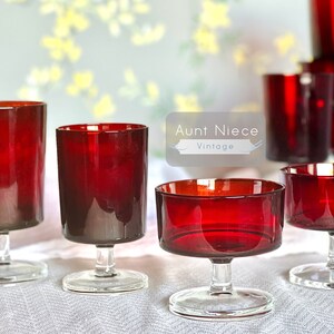 May include: Set of six vintage red glass stemmed drinking glasses. The glasses are in various shapes and sizes, including a tall cylinder, a shorter cylinder, and a coupe shape. The glasses are all clear and have a red tint.
