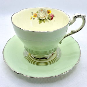 May include: A vintage teacup and saucer set with a delicate floral design. The cup is white with a green band and a silver rim. The saucer is pale green with a silver rim.