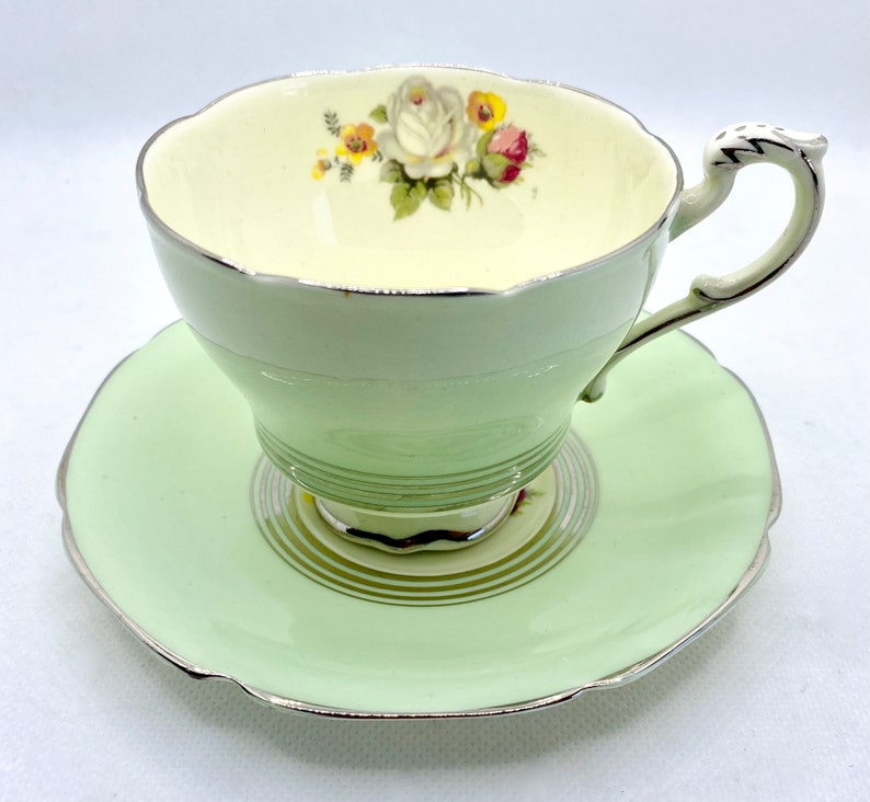 May include: A vintage teacup and saucer set with a delicate floral design. The cup is white with a green band and a silver rim. The saucer is pale green with a silver rim.