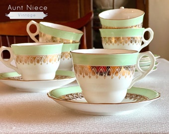 May include: Set of six vintage teacups and saucers with a green and gold design. The cups and saucers are white with a green band and gold trim. The gold trim features a repeating pattern of stylised leaves and flowers.