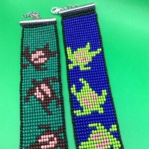 May include: Two beaded bracelets with a clasp closure. One bracelet is green with brown and pink turtles, the other is blue with green and pink turtles.