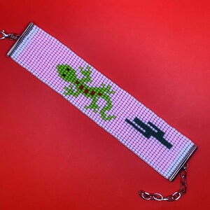 May include: A pink beaded bracelet with a green lizard and a cactus design. The bracelet has a silver clasp and chain.