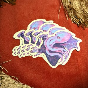 May include: A stack of stickers featuring a Cthulhu design. The stickers have a white border and depict the creature in shades of purple, blue, and pink. The stickers are on a red velvet surface with gold fringe.