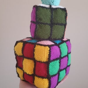 Könnte beinhalten: Ein farbenfrohes gehäkeltes Rubik's Cube Spielzeug mit einem kleineren grünen gehäkelten Würfel oben drauf. Der größere Würfel hat rote, gelbe, grüne, violette und schwarze Quadrate.