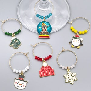 May include: A collection of Christmas-themed wine charms. Each charm features a gold-colored ring with a festive pendant, including a snowman, a mitten, a penguin, a snow globe with a bear, a red hat, and a snowflake. The charms are adorned with colorful beads.