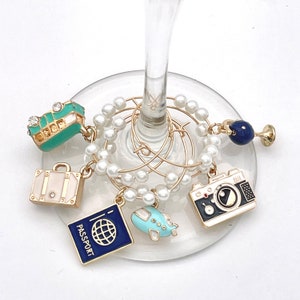 May include: Wine glass charm set with travel-themed charms. The set includes charms shaped like a suitcase, passport, airplane, camera, and a teal bus. The charms are gold-toned and hang from a gold wire ring with faux pearls.
