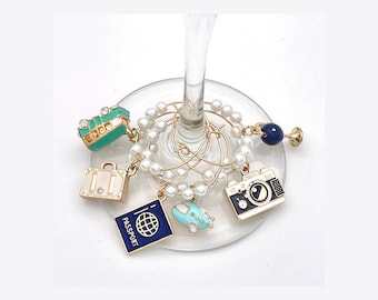 May include: A set of circular wine glass charms featuring white faux pearls and gold-tone metal rings. The charms include a teal trolley, a beige suitcase, a dark blue passport, a light blue airplane, a black camera, and a dark blue bead.
