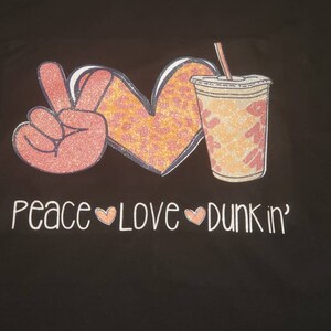 May include: Black t-shirt with a pink peace sign, a heart with orange and pink leopard print, and a pink and orange iced coffee cup with a straw. The text "peace love dunkin'" is printed below the images.