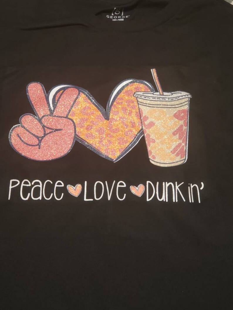 May include: Black t-shirt with a pink peace sign, a heart with orange and pink leopard print, and a pink and orange iced coffee cup with a straw. The text "peace love dunkin'" is printed below the images.
