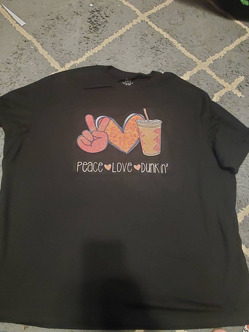 May include: Black t-shirt with a peace sign, heart, and a cup of coffee with the text "Peace Love Dunkin'" printed on it.