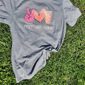 May include: Gray t-shirt with a pink peace sign, a pink heart with a leopard print, and a brown and white coffee cup with a straw. The text "Peace Love Dunkin'" is printed below the graphic.