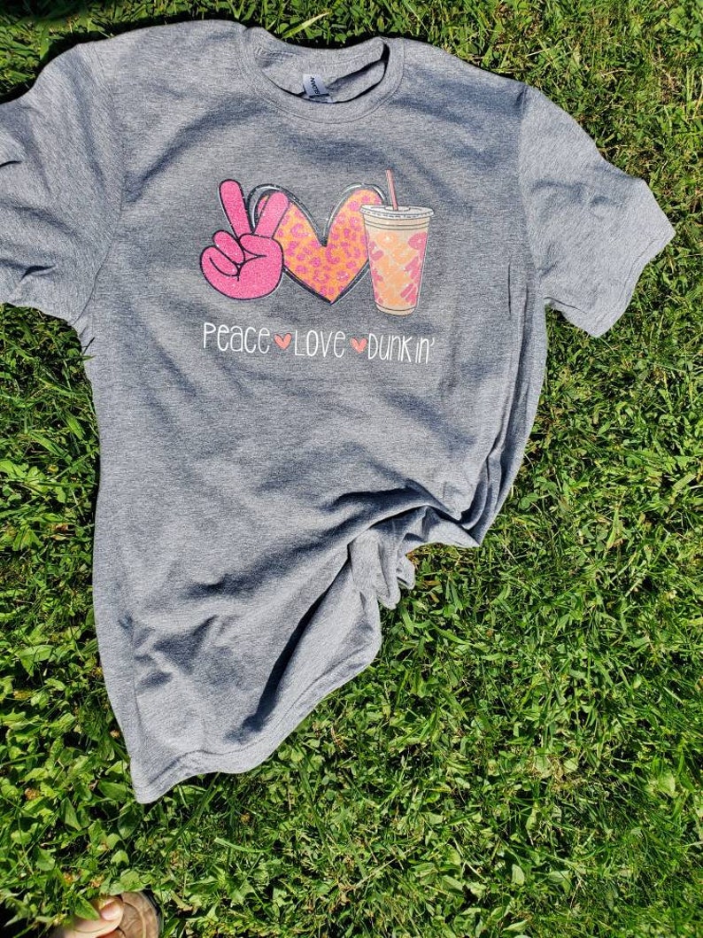 May include: Gray t-shirt with a pink peace sign, a pink heart with a leopard print, and a brown and white coffee cup with a straw. The text "Peace Love Dunkin'" is printed below the graphic.