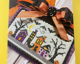 May include: A cross-stitch pattern card with a black and white image of a spooky Halloween village with houses, trees, and bats. The card has a black background with white text that reads "Primrose Cottage Stitches" and "Witchy Way".