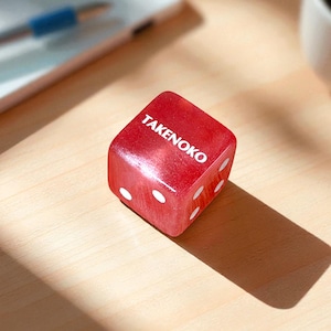 May include: A red TAKENOKO die with white dots, resting on a wooden surface. The die is a cube shape, with the word TAKENOKO printed in white on one side. The dots are arranged in the standard pattern for a six-sided die.