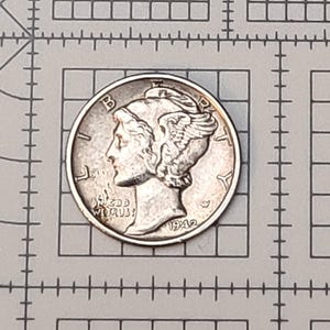 May include: A silver Mercury dime coin, featuring a profile of a woman with a winged cap, is centered on a grid background. The coin's details are visible, including the inscription "LIBERTY" and the date "1942".