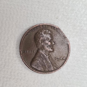 Puede incluir: Una moneda de cobre Lincoln de 1944 con un borde dañado. El anverso presenta un perfil de Abraham Lincoln, la palabra "LIBERTY" y la fecha. También son visibles las palabras "IN GOD WE TRUST".