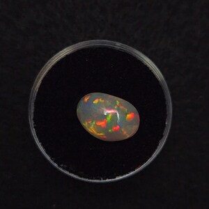 Puede incluir: Una piedra preciosa de ópalo ovalada y pulida con juego de colores, incluyendo destellos rojos, naranjas y verdes. El ópalo está colocado sobre un fondo negro dentro de una vitrina circular transparente. La piedra preciosa mide aproximadamente 2 cm de largo.