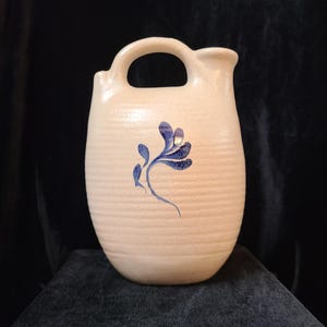 Puede incluir: Una jarra de cerámica de color crema con un diseño floral azul. La jarra tiene un asa y un pico.