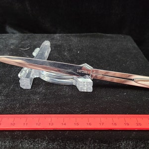 May include: A silver letter opener with the word "DIETZGEN" engraved on the blade. The letter opener is resting on a clear plastic stand.