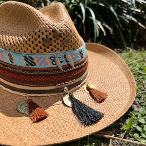 May include: A straw cowboy hat with a brown band and multiple layers of trim, including leather, beads, and tassels. The hat has a woven texture and is decorated with metal accents.