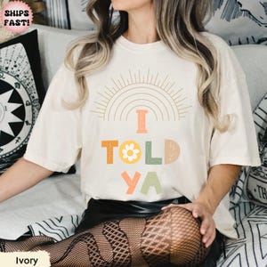May include: Ivory-colored t-shirt with the text "I TOLD YA" in colorful, playful letters. The design includes a sun and rainbow graphic. The shirt is a relaxed fit.