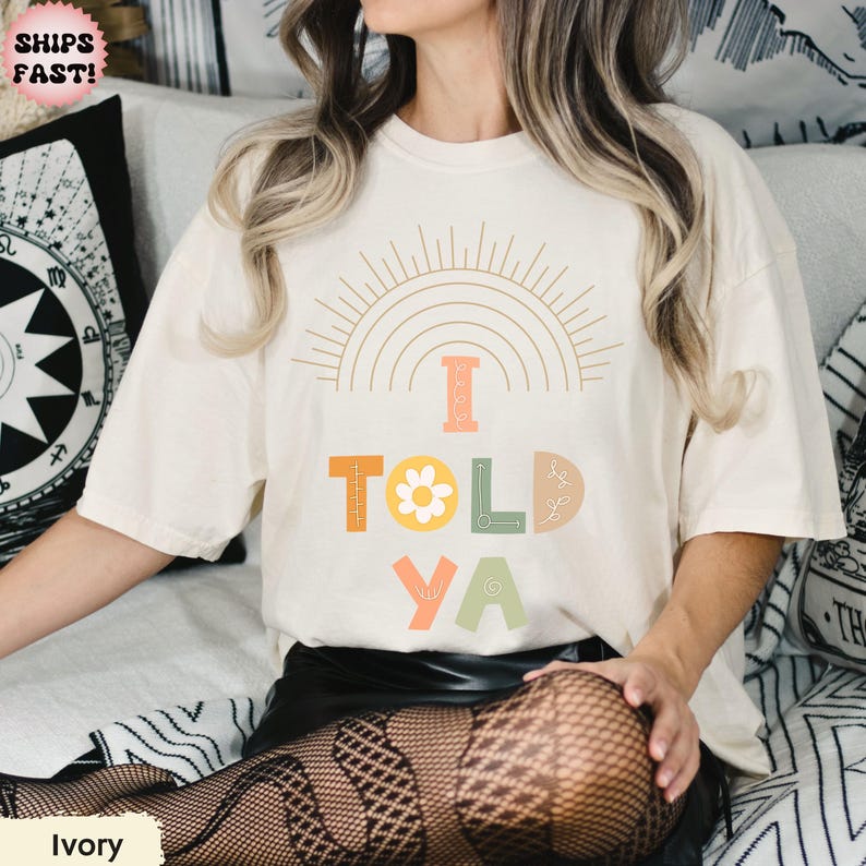 May include: Ivory-colored t-shirt with the text "I TOLD YA" in colorful, playful letters. The design includes a sun and rainbow graphic. The shirt is a relaxed fit.