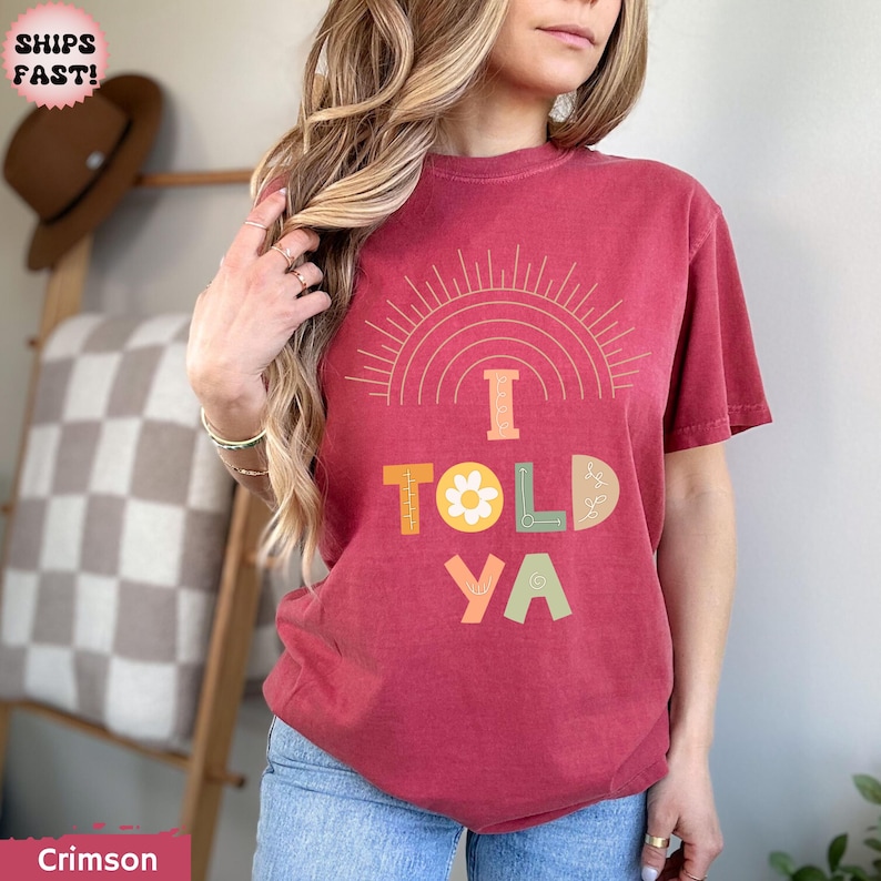 May include: Crimson-colored t-shirt with the text "I TOLD YA" in colorful, playful letters. The design includes a sunburst and a flower. The shirt is a comfortable, casual style.