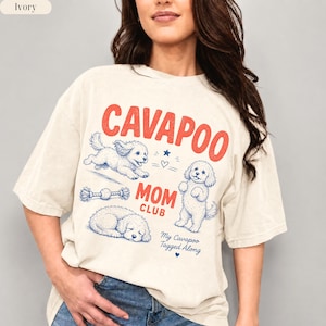 May include: An ivory t-shirt featuring red text that reads CAVAPOO MOM CLUB and blue illustrations of Cavapoo dogs in various poses, including running, standing, and lying down, along with a rope toy and the text My Cavapoo Tagged Along.