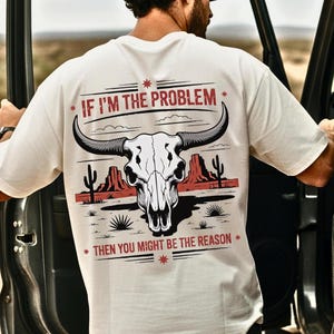 Puede incluir: Camiseta color crema con un gráfico de una calavera de toro, un paisaje desértico y el texto "IF I'M THE PROBLEM THEN YOU MIGHT BE THE REASON." El diseño presenta acentos rojos y negros.