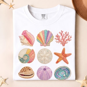 Puede incluir: Camiseta blanca con diseños de conchas bordadas de colores. La camiseta incluye una estrella de mar, coral y diferentes tipos de conchas. La etiqueta dice "Comfort Colors".