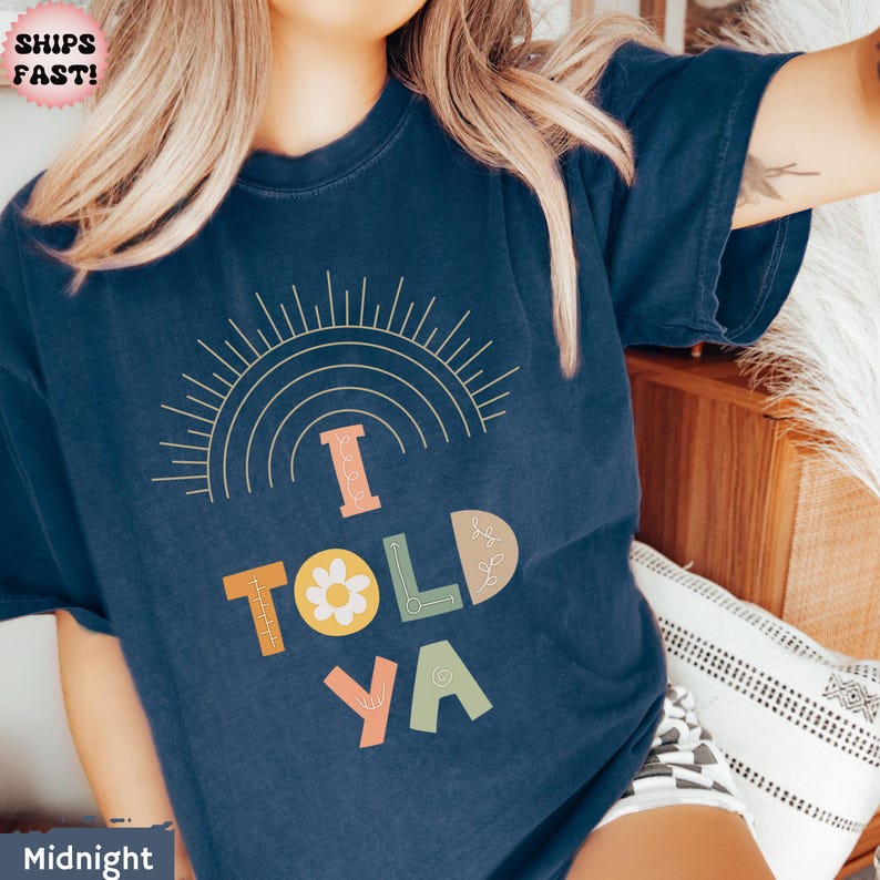 May include: Midnight blue t-shirt with the text "I TOLD YA" in a colorful, retro font. The design includes a sunburst and rainbow graphic above the text. The shirt is a comfortable, casual style.