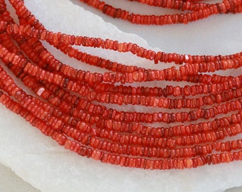 Puede incluir: Una hebra de cuentas de coral rojo, de aproximadamente 4 mm de diámetro, expuesta sobre una superficie blanca. Las cuentas son uniformes en tamaño y forma, creando un aspecto liso y consistente.