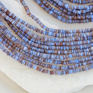 Può includere: Un filo di perline di vetro blu e marrone, di circa 4 mm di dimensione. Le perline sono disposte in un mucchio sciolto su una superficie bianca.