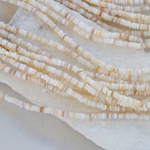 Peut inclure: Un brin de perles de coquillage blanches et brunes, d'environ 4 mm de diamètre. Les perles sont disposées en un tas lâche sur une surface blanche.