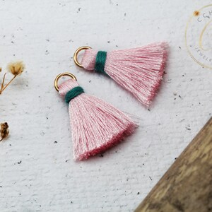 Op de afbeelding: Twee roze kwastbedels met gouden springringen en groene trim. De kwasten zijn gemaakt van zacht, pluizig garen en zijn perfect om een ​​toets van whimsy toe te voegen aan sieraden of knutselwerk.
