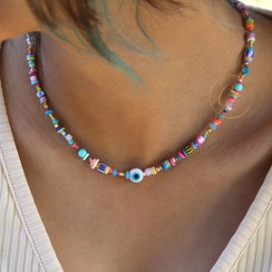 Puede incluir: Un collar de cuentas de colores con un dije de ojo turco azul y blanco. El collar está hecho con una variedad de cuentas pequeñas y coloridas, incluyendo rosa, azul, amarillo, verde y naranja.