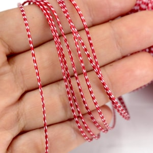 Puede incluir: Primer plano de hilo rojo y blanco sostenido en una mano. El hilo está bien enrollado y tiene un patrón repetitivo de rojo y blanco. Un carrete del mismo hilo es visible en el fondo.