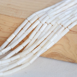 Pu&ograve; includere: Una collana di perline bianche di conchiglia, ogni perlina ha una forma cilindrica. Le perline sono disposte in fila su una superficie di legno marrone chiaro.
