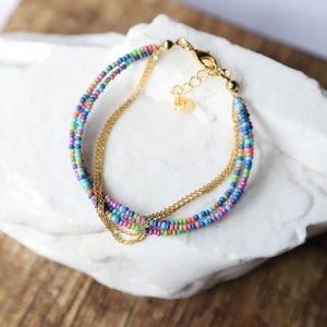 Può includere: Un braccialetto multifilo con perline colorate e una catena dorata. Il braccialetto è realizzato con piccole perline rotonde in tonalità di blu, verde, rosa e arancio. La catena dorata è intrecciata con le perline, creando un effetto a strati.